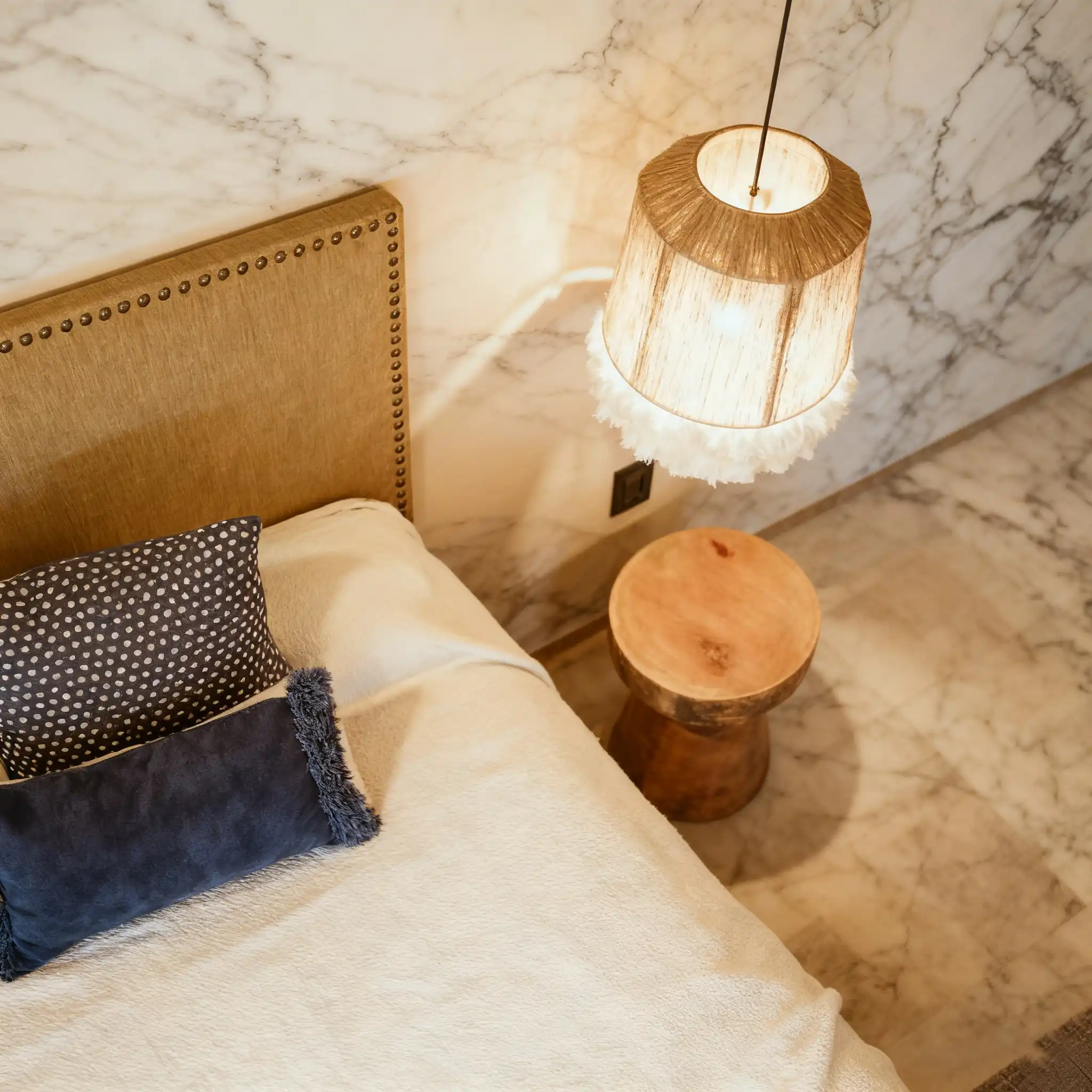A bedroom corner with a tufted headboard, patterned pillows, a wooden stump side table, and a pendant light with a fringed lampshade, against a marble wall.